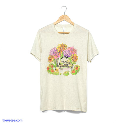 Natural oatmeal colored tee shirt with Chicory character centered sitting on bench with paintbrush leaned on bench surrounded by colorful flowers and foliage