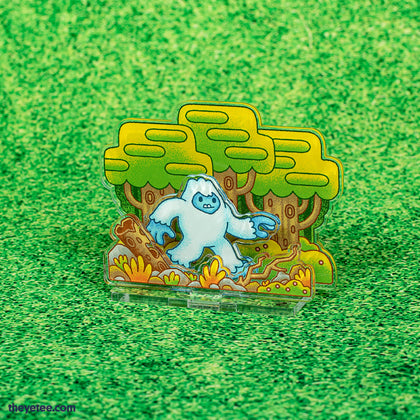1 x 4" x 6" Flat Acrylic Stand of the Yetee walking through a forest. Foreground consists of rocks and small bushes and the background is three tall trees. A small acrylic hand-held videogame is included. 